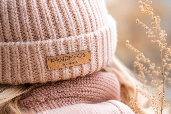 Étiquette rectangulaire beige en similicuir « Handmade by Bine » sur un bonnet rose en maille.